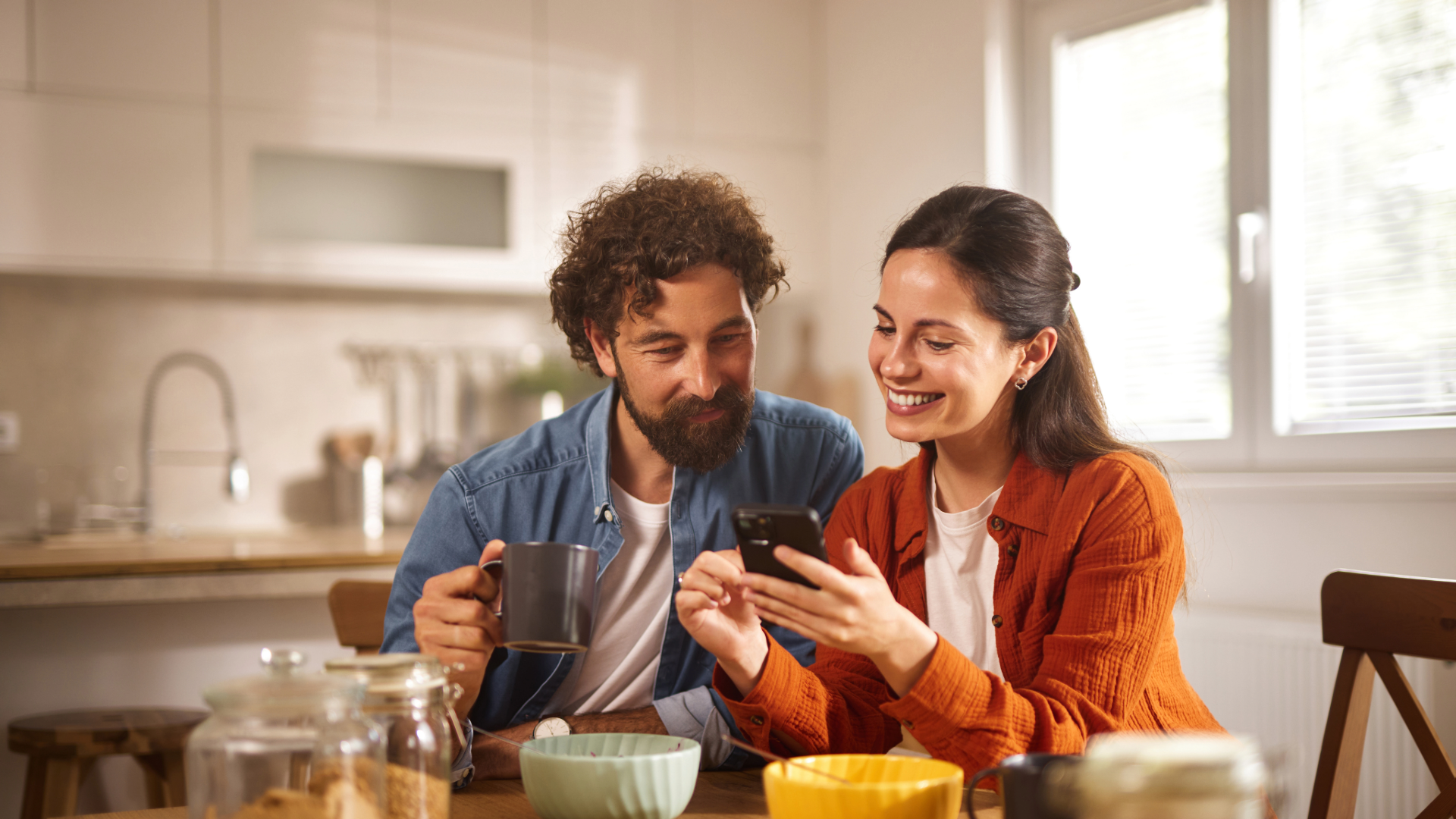 Untitled - October 26, 2026 at 14.53.15 A couple sits at a wooden table in their home office, smiling and sharing a moment while looking at a smartphone.
